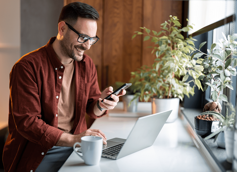 Man smiling, using phone and laptop indoors.